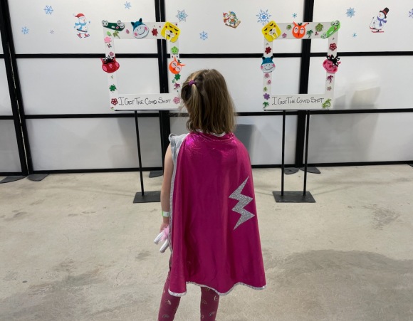 Beatrix, in a pink superhero cape with a silver lightning bolt on it, looking at/deciding between two "I got my covid shot" photo frames for taking an "after shot" photo