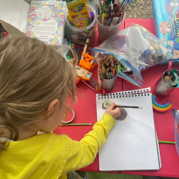 Overhead photo of beatrix drawing a kiwi in a sketchbook, surrounded by art supplies